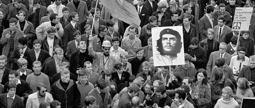 Schwarz-Weiss-Fotografie einer grossen Menschenmenge bei einer Demonstration in den 1960er-Jahren. In der Mitte tragen Teilnehmende ein grosses Porträt von Che Guevara und eine Fahne mit Stern. Rechts im Bild ist ein Schild mit der Aufschrift „Keine Unterstützung für die US-Politik in Vietnam!“ zu sehen.