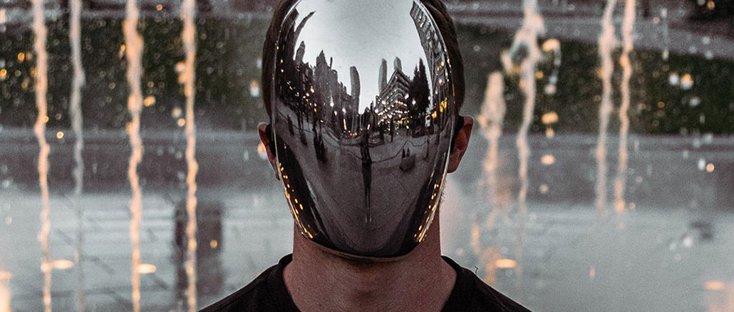 Person mit einer spiegelglatten, metallisch glänzenden Maske ohne erkennbare Gesichtszüge, in der sich die umliegende Stadtlandschaft und Passanten spiegeln. Im Hintergrund spritzen Wasserfontänen in die Höhe. Die Szene wirkt futuristisch und anonymisierend.