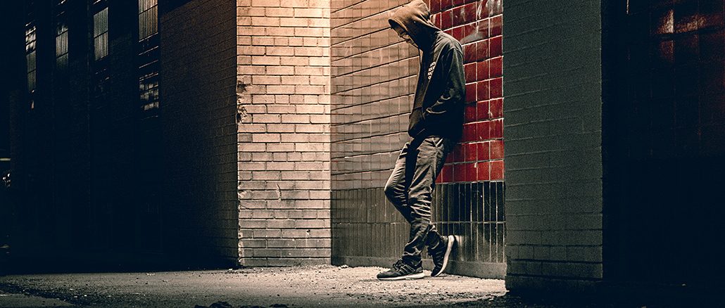 Eine Person im Kapuzenpullover lehnt nachts an einer Hauswand mit roten und weissen Kacheln. Der Kopf ist gesenkt, die Hände in den Taschen, das Gesicht bleibt im Schatten verborgen. Die Szene ist von einer Strassenlaterne beleuchtet und wirkt einsam und düster.