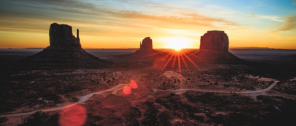 Sonnenaufgang über dem Monument Valley: Drei markante rote Sandstein-Buttes ragen aus der weiten, kargen Wüstenlandschaft empor, während die Sonne am Horizont aufgeht und goldene Strahlen über Himmel und Erde legt.