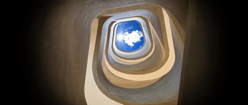 Blick von unten durch ein spiralförmiges, organisch geschwungenes Treppenhaus nach oben zum Himmel. In der Mitte ist ein rechteckiger Ausschnitt mit blauem Himmel und einer weissen Wolke sichtbar, durch den Sonnenlicht hereinfällt.