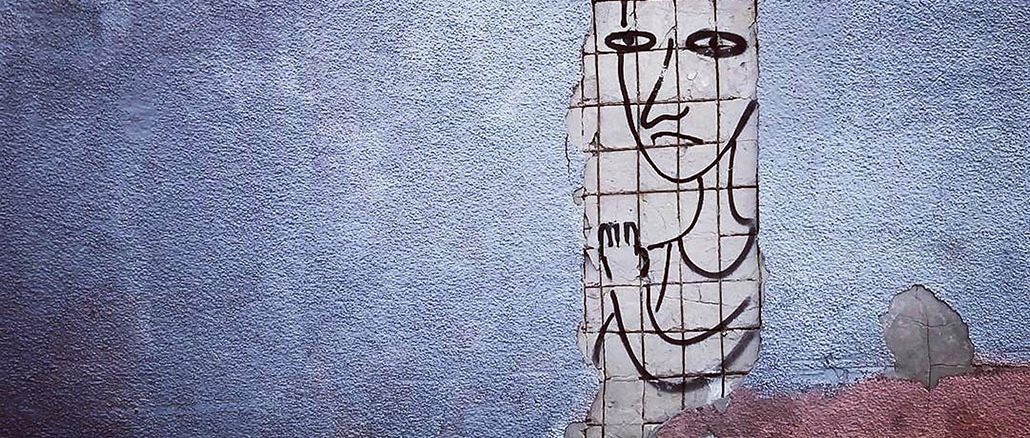 Auf einer verblassten, teils abbröckelnden Mauer ist ein einfaches, schwarz umrandetes Gesicht im Stil einer Strichzeichnung zu sehen, auf den freigelegten Fliesen darunter gezeichnet. Die Figur wirkt schemenhaft und melancholisch, als ob sie hinter der Wand hervorblickt.