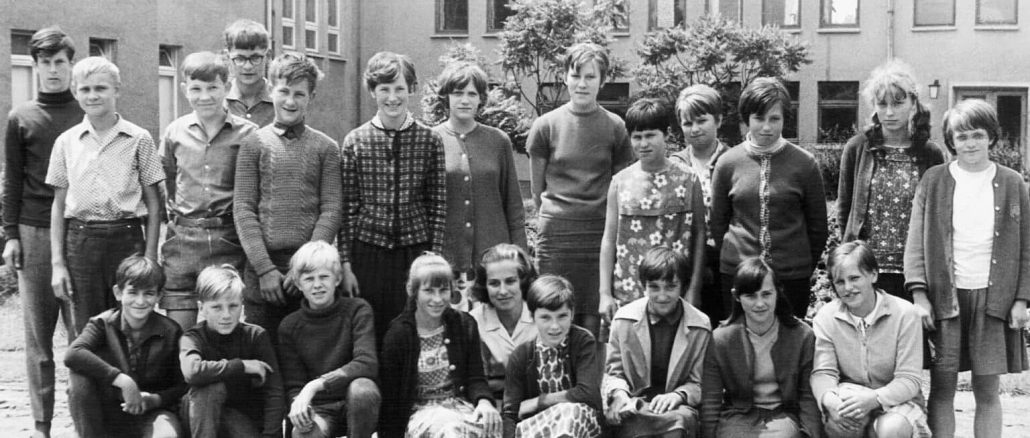 Schwarz-Weiss-Fotografie einer Schulklasse aus den 1960er-Jahren: Eine Gruppe von Jungen und Mädchen posiert in zwei Reihen vor einem Schulgebäude. Vorne sitzen einige Kinder auf dem Boden, dahinter stehen die übrigen. Kleidung und Frisuren spiegeln den Stil der Zeit wider, die Stimmung wirkt formell, aber freundlich.