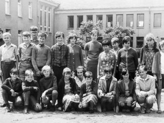 Schwarz-Weiss-Fotografie einer Schulklasse aus den 1960er-Jahren: Eine Gruppe von Jungen und Mädchen posiert in zwei Reihen vor einem Schulgebäude. Vorne sitzen einige Kinder auf dem Boden, dahinter stehen die übrigen. Kleidung und Frisuren spiegeln den Stil der Zeit wider, die Stimmung wirkt formell, aber freundlich.
