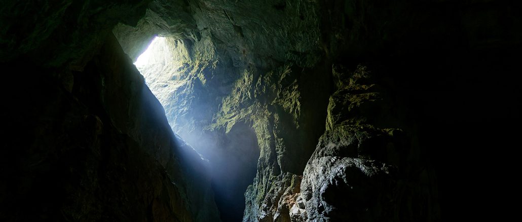 Ein schwach beleuchteter Höhlenraum: Grobe, unregelmäßige Felswände und eine gewölbte Decke, nasses Gestein spiegelt leicht das Licht. In der Ferne ist ein Spalt oder Tunnel sichtbar, durch den schwaches, kaltes Licht einfällt. Atmosphäre: klaustrophobisch, geheimnisvoll.