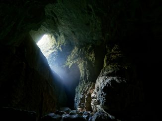 Ein schwach beleuchteter Höhlenraum: Grobe, unregelmäßige Felswände und eine gewölbte Decke, nasses Gestein spiegelt leicht das Licht. In der Ferne ist ein Spalt oder Tunnel sichtbar, durch den schwaches, kaltes Licht einfällt. Atmosphäre: klaustrophobisch, geheimnisvoll.