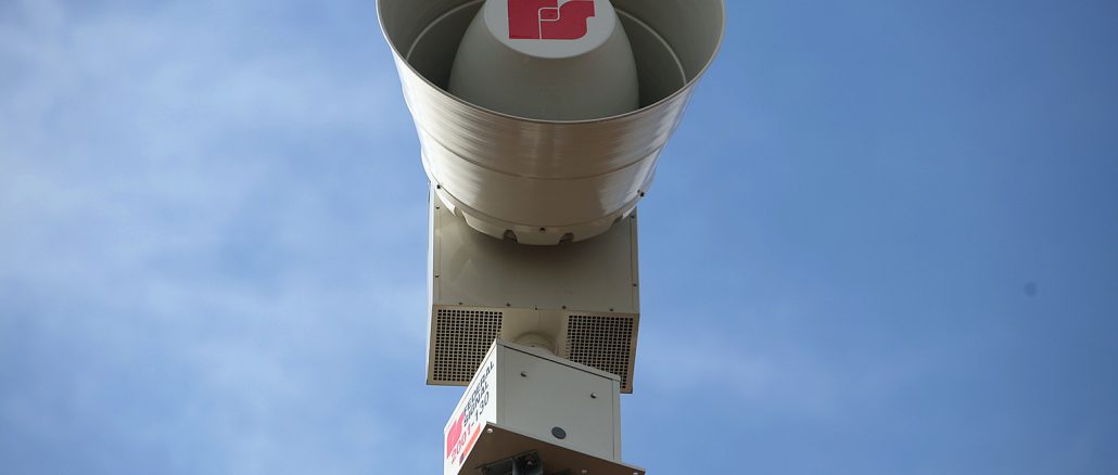 Aussenaufnahme einer Warnsirene auf einem Holzpfosten vor blauem Himmel.