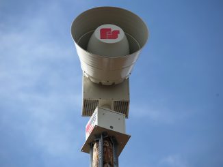 Aussenaufnahme einer Warnsirene auf einem Holzpfosten vor blauem Himmel.