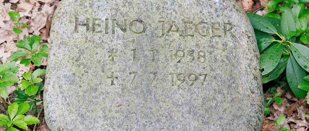 Grabstein aus grauem Granit mit der eingravierten Inschrift „HEINO JAEGER * 1.1.1938 † 7.7.1997“, umgeben von grünem Laub und fallengelassenem Laub auf dem Boden.