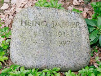 Grabstein aus grauem Granit mit der eingravierten Inschrift „HEINO JAEGER * 1.1.1938 † 7.7.1997“, umgeben von grünem Laub und fallengelassenem Laub auf dem Boden.