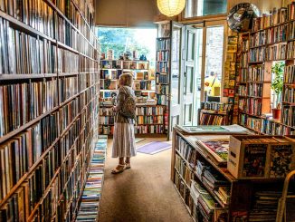 Eine Frau steht in einem kleinen, lichtdurchfluteten Antiquariat zwischen hohen Bücherregalen und betrachtet die Buchrücken. Durch die geöffnete Tür fällt Tageslicht ins Geschäft, das von Regalen, Comics und Stapeln alter Bücher gefüllt ist.