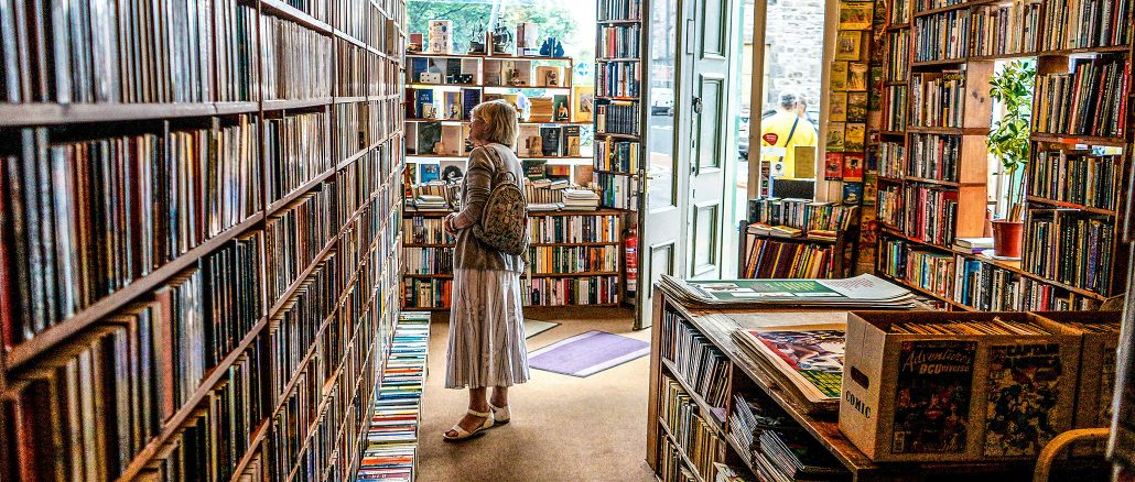 Eine Frau steht in einem kleinen, lichtdurchfluteten Antiquariat zwischen hohen Bücherregalen und betrachtet die Buchrücken. Durch die geöffnete Tür fällt Tageslicht ins Geschäft, das von Regalen, Comics und Stapeln alter Bücher gefüllt ist.