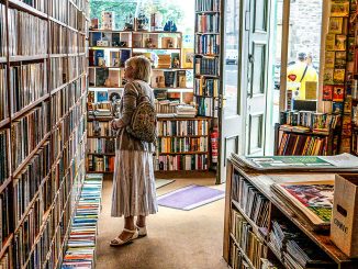 Eine Frau steht in einem kleinen, lichtdurchfluteten Antiquariat zwischen hohen Bücherregalen und betrachtet die Buchrücken. Durch die geöffnete Tür fällt Tageslicht ins Geschäft, das von Regalen, Comics und Stapeln alter Bücher gefüllt ist.