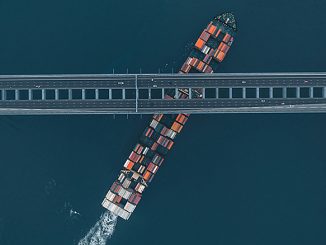 Containerfrachter mit bunt gestapelten Containern fährt schräg unter einer Brücke hindurch; Luftaufnahme, die die enge Passage und globale Transportinfrastruktur zeigt.