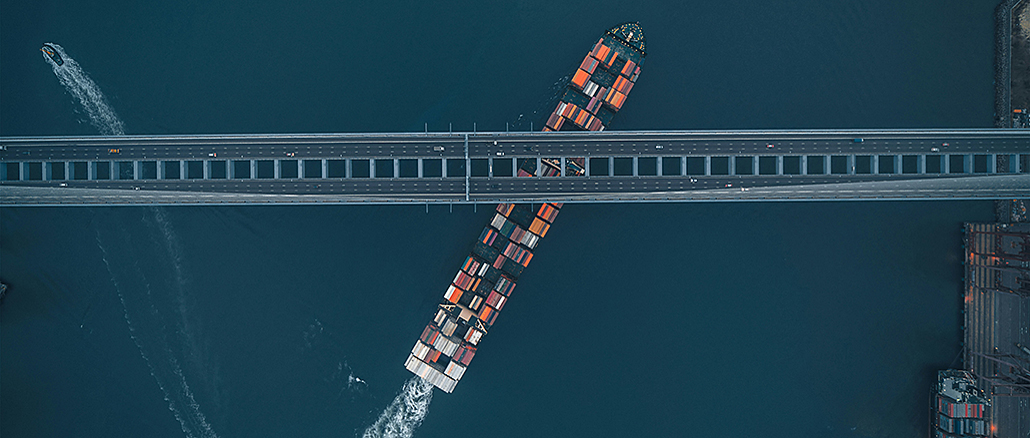 Containerfrachter mit bunt gestapelten Containern fährt schräg unter einer Brücke hindurch; Luftaufnahme, die die enge Passage und globale Transportinfrastruktur zeigt.