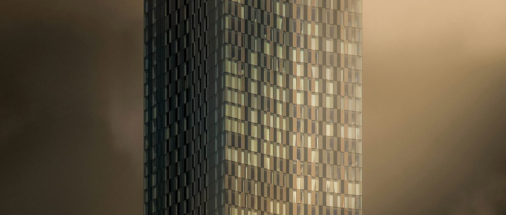 Hochhausfassade mit vielen kleinen Fensterquadraten, teilweise reflektierend in warmen Gold- und Bronzetönen, vom unteren Bildrand her aufsteigender dichter Nebel oder Dampf, der das Gebäude halb verdeckt; Stimmung düster-mystisch.