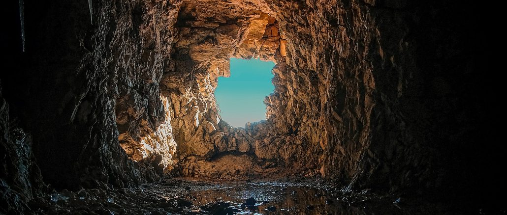 Blick aus dem Inneren einer dunklen, felsigen Höhle durch eine unregelmässige Öffnung nach draußen; warmes Licht beleuchtet die zerklüfteten Steinwände, auf dem Höhlenboden spiegelt sich Wasser, dahinter ist ein klarer, blauer Himmel sichtbar.