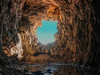 Blick aus dem Inneren einer dunklen, felsigen Höhle durch eine unregelmässige Öffnung nach draußen; warmes Licht beleuchtet die zerklüfteten Steinwände, auf dem Höhlenboden spiegelt sich Wasser, dahinter ist ein klarer, blauer Himmel sichtbar.