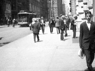 Schwarzweissfotografie einer belebten New Yorker Strasse im frühen 20. Jahrhundert: Männer in Anzügen und Hüten gehen über den Bürgersteig, im Hintergrund fahren eine Strassenbahn und Pferdefuhrwerke; im Vordergrund kommt ein Mann im dunklen Anzug mit Hut auf die Kamera zu.