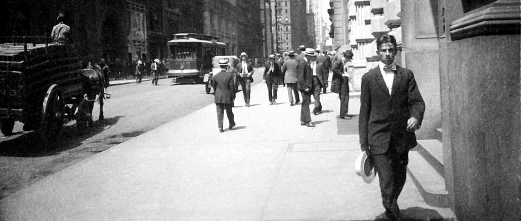 Schwarzweissfotografie einer belebten New Yorker Strasse im frühen 20. Jahrhundert: Männer in Anzügen und Hüten gehen über den Bürgersteig, im Hintergrund fahren eine Strassenbahn und Pferdefuhrwerke; im Vordergrund kommt ein Mann im dunklen Anzug mit Hut auf die Kamera zu.