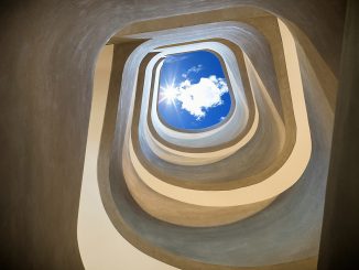 Blick von unten durch ein spiralförmiges, organisch geschwungenes Treppenhaus nach oben zum Himmel. In der Mitte ist ein rechteckiger Ausschnitt mit blauem Himmel und einer weissen Wolke sichtbar, durch den Sonnenlicht hereinfällt.