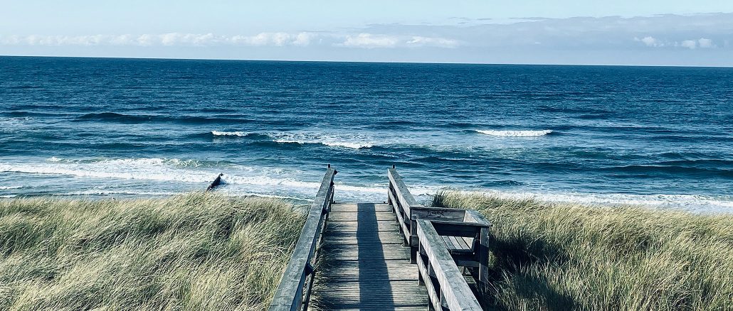 Holzsteg durch Dünengras zum Strand auf Sylt, dahinter die weite Nordsee unter klarem Himmel.