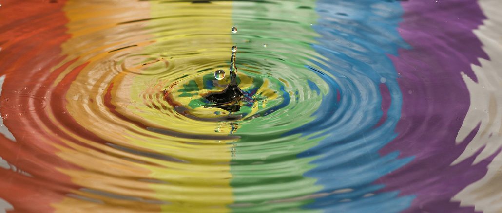 Wassertropfen fällt in eine farbig schillernde Oberfläche, die in Regenbogenfarben leuchtet; konzentrische Kreise breiten sich von der Einschlagstelle aus, während ein kleiner Tropfen nach oben spritzt.