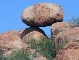 Grosser rundlicher Felsblock liegt auf mehreren rotbraunen Felsen unter blauem Himmel.