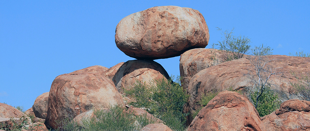 Grosser rundlicher Felsblock liegt auf mehreren rotbraunen Felsen unter blauem Himmel.